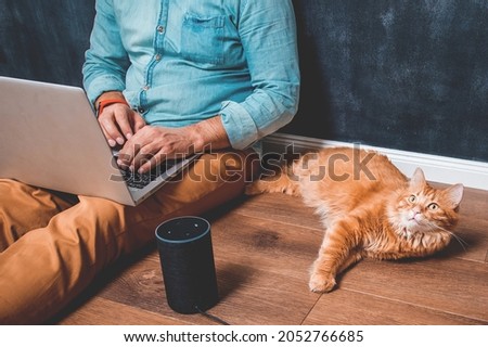 Workspace at home office. Man with laptop sitting on wooden floor. Pet cat lying near alexa speaker. Man working at the computer. Playing music from voice smart control.
