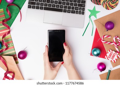 Workspace with laptop and wrapping paper, top view. Flat Lay Christmas or Party Background with ribbons, candy canes, baubles, Star tag and decorations. Female hands holdings phone - Powered by Shutterstock