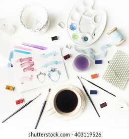 Workspace. Logo Painted With Watercolor On Paper, Spool With Blue Ribbon, Sketchbook, Pot, Palette, Brushes And Black Coffee Cup Isolated On White Background. Flat Lay, Overhead View, Top View