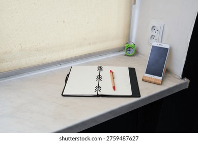 A workspace setup featuring an open lined notebook, a pen, a small green alarm clock, and a smartphone on a wooden stand, placed on a windowsill