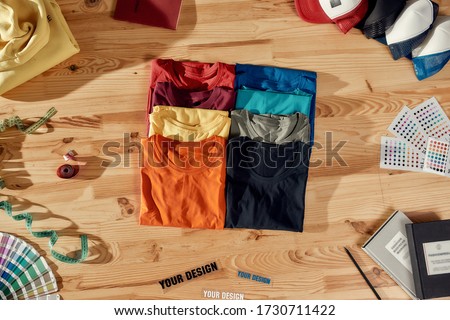 Workspace. Top view of neatly folded custom t shirts and arranged baseball caps. Samples of fabric, stickers with text, tape measure and notebooks lying on a wooden background. Horizontal shot