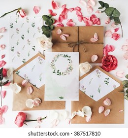 Workspace. Wedding invitation cards, craft envelopes, pink and red roses and green leaves on white background. Overhead view. Flat lay, top view - Shutterstock ID 387517810