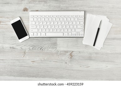 Workstation At A White Rustic Wooden Desk With A Mobile Phone, Computer Keyboard And Notepad And Pen Arranged In A Line Above Copy Space, Overhead View