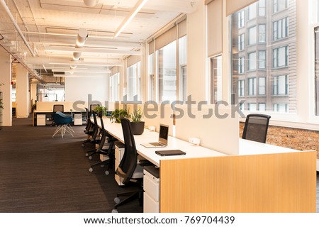 Workstations sprawl across a modern,  open concept office building. Natural light fills the room from large windows along the walls.