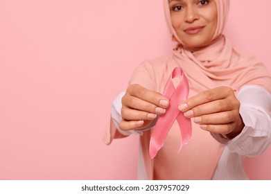 World Cancer Day concept. Focus on pink Breast Cancer Awareness ribbon in the manicured hands of blurred beautiful friendly Arab Muslim woman in pink hijab headscarf on pink background with copy space - Shutterstock ID 2037957029