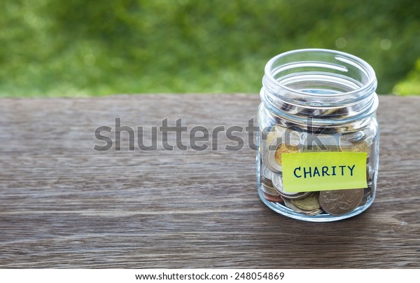 World coins in money\
glass jar with CHARITY word label place on natural wood table,\
blank space for text