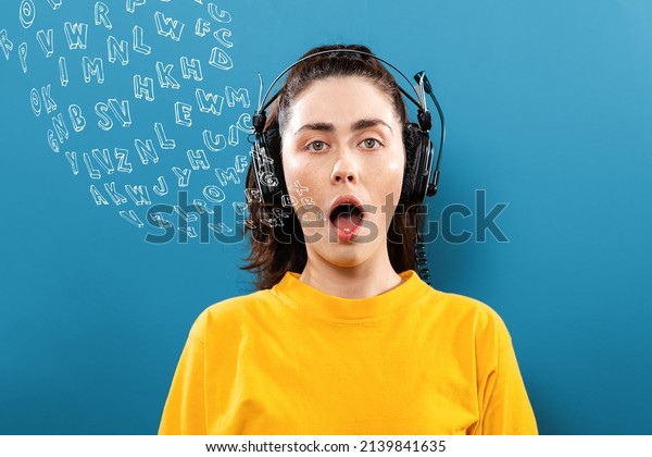 World English Language Day.\
Portrait of a young woman wearing headphones, with her mouth open.\
Blue background with letters. The concept of learning foreign\
languages.