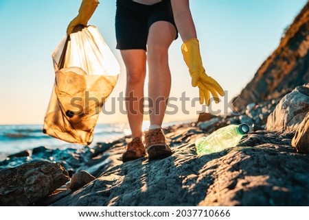 World environment day. An activist picks up a plastic bottle on the beach. Close up. In the background, the sea and the sunset. Bottom view. The concept of cleaning the coastal zone.