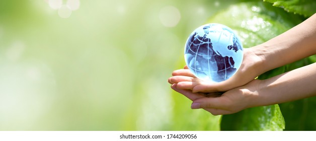 World Environment Day or Earth Day concept. Blue glass globe in woman hands on blurred green leaves banner background. Saving planet, save, protect clean nature and ecology, sustainable lifestyle. 