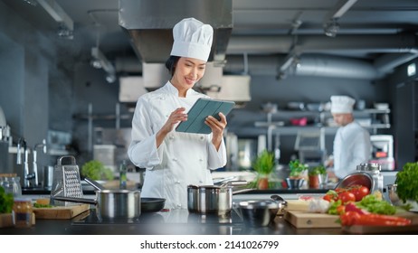 World Famous Restaurant: Asian Female Chef Cooking Delicious and Authentic Food, Uses Digital Tablet Computer While Working in a Modern Professional Kitchen. Preparing gourmet organic Dishes - Shutterstock ID 2141026799