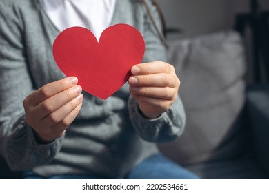 World Heart Day, World Health Day, CSR Responsibility, Adoption Foster Care Home, All Lives Matter, No To Racism Concept. Close-up Of Young Female Holding Small Red Paper Heart Sitting On Comfy Couch