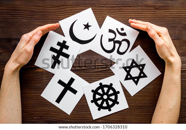 World religions concept. Hands hugs Christianity,\
Catholicism, Buddhism, Judaism, Islam symbols on wooden background\
top view