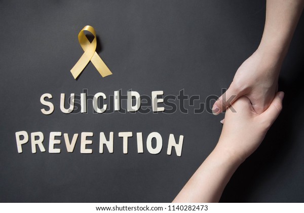 World suicide prevention day - Holding hands for\
helping and supporting depressed woman with yellow ribbon awareness\
and SUICIDE PREVENTION wooden word on black background. Mental\
health care concept.
