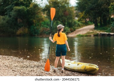 World Tourism Day. A young happy woman stands with an oar in her hand near the kayak. Back view. The concept of kayaking and outdoor activities