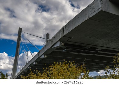 A worm's-eye view of the concrete underside and cable pylon of a modern cable-stayed bridge against a dramatic blue sky with white clouds.