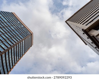 Worm's-eye view looking straight up between two modern skyscrapers toward a cloudy sky during the day. The perspective represents rapid urban growth, ambitious development, future cities, and corporate scale.