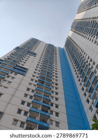 A worm's-eye view looking up at two tall, modern apartment buildings against a clear sky. The buildings are primarily white with prominent blue horizontal balcony railings and a vertical blue stripe on the facade of the building on the right. Numerous windows and balconies are visible across many floors.