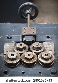 Worn industrial roller assembly with multiple metal rollers, hex nuts, washers, and mounting plates fixed to painted machinery base, showing grease, rust, texture, and signs of long-term service