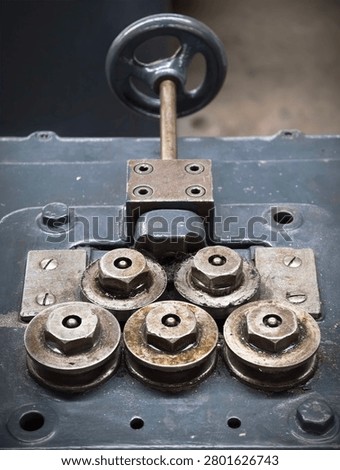Worn industrial roller assembly with multiple metal rollers, hex nuts, washers, and mounting plates fixed to painted machinery base, showing grease, rust, texture, and signs of long-term service