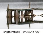 A worn and textured wooden plank wharf. The boards are grey and one horizontal green one with smooth water in the background.The water has lights reflecting on it. The pier is small for pleasure craft