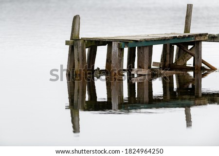 A worn and textured wooden plank wharf. The boards are grey and one horizontal green one with smooth water in the background.The water has lights reflecting on it. The pier is small for pleasure craft