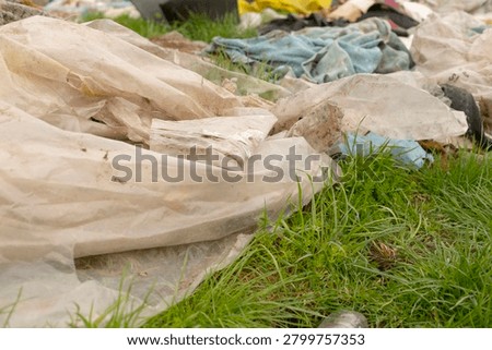 Worn translucent plastic sheets and dirty fabric pieces strewn across a patch of green grass, creating a messy outdoor scene. The foreground shows crumpled