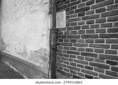 A worn wall meets a brick structure at a corner. A metal pipe runs down the wall. The scene is in black and white. The setting shows signs of age and wear.