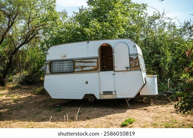 A worn-out white caravan, missing its wheels, sits abandoned in a rural landscape. Its exterior is weathered and stained, blending into the quiet and desolate surroundings of the countryside.