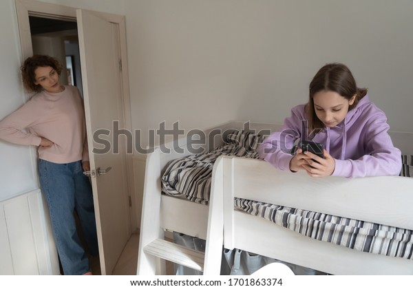 Worried adult parent mom coming to teenage child\
bedroom unhappy about technology addicted kid using phone, obsessed\
with mobile social media apps. Teens gadgets overuse and parental\
control concept.