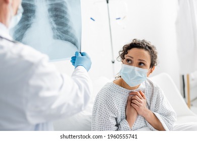 Worried African American Woman In Medical Mask Near Radiologist In Latex Glove Holding Lungs X-ray On Blurred Foreground