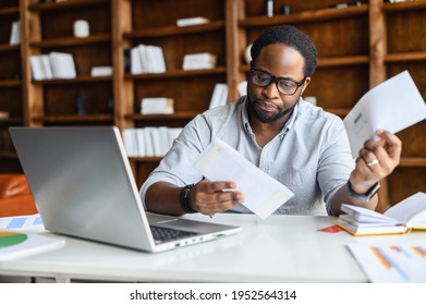 Worried African-American looking through correspondence, a guy holds envelopes in hands, got fine by post, concentrated man sitting at the desk with a laptop in contemporary office