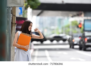 Worried Asian Businesswoman Looking At Her Watch While Waiting Cab In A City. Beautiful Female Is Calling And Hailing A Cab In A City. Traffic Jam, Rush Hours, Hurry Up, Time, Late. Copy Space. 