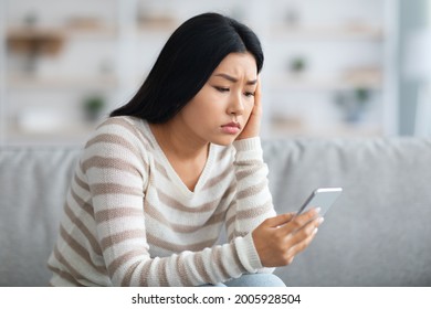 Worried Asian Lady Looking At Smartphone Screen While Sitting On Sofa At Home, Frustrated Korean Woman Holding Mobile Phone, Got Bad News Or Reading Unplesant Message, Closeup Shot, Free Space