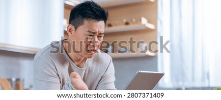 Worried Asian Man Using Tablet In Kitchen, Facing Technology Issue, Stressful Situation