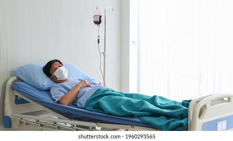 Worried Asian middled-aged male patient getting blood transfusion in private room at hospital ward. Senior man wears a white protective facemask for personal hygiene concept