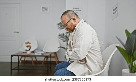 Worried, attractive middle-aged caucasian man suffering serious stomachache while sitting uneasily on a chair in a medically emphasized waiting room, his stressed expression hinting sickness. - Powered by Shutterstock