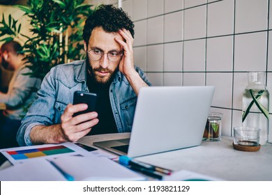 Worried Bearded Young Man Reading Sms Message With Bad Notification On Modern Smartphone Sitting At Laptop Computer In Cafe.Frustrated And Upset Hipster Guy Watching Shocked Video In Blog On Cellular