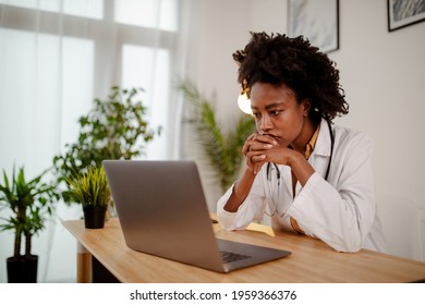 Worried Black Female Doctor Working On Computer At Her Desk.