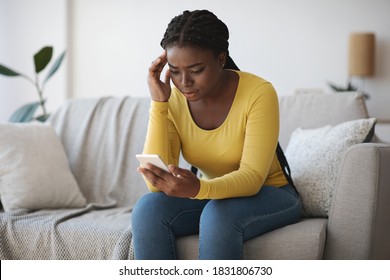 Worried Black Lady Looking At Smartphone Screen, Sitting On Couch At Home, Concerned African American Woman Waiting For Important Call, Reading Unpleasant Sms, Spending Time With Phone In Living Room