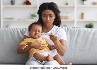 Worried Black Mother Checking Temperature Of Her Crying Infant Baby At Home, African Mom Looking At Thermometer While Sitting On Couch In Living Room, Little Toddler Child Having Fever, Copy Space