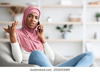 Worried Black Muslim Woman In Hijab Talking On Cellphone At Home, Frustrated Islamic Lady Having Unpleasant Conversation While Sitting On Chair In Living Room, Emotionally Reacting To Bad News - Powered by Shutterstock