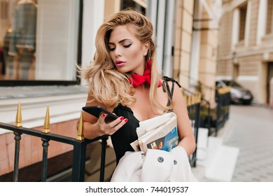 Worried Blonde Female Model Checking Social Networks While Standing Beside Restaurant Alone. Attractive Sad Woman In Elegant Outfit Typing Message On Phone Holding Coat In Hand.