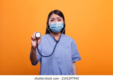 Worried Concerned Healthcare Clinic Asian Nurse Wearing Protective Facemask While Holding Stethoscope. Distressed Caretaker Wearing Virus Protection Mask While Holding Medical Instrument.