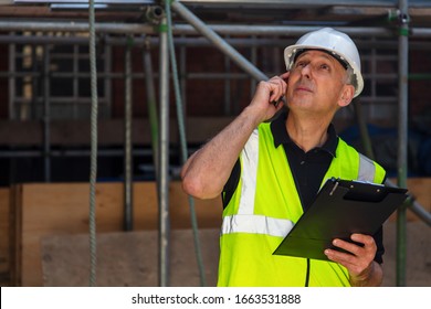 Worried Or Concerned Male Builder Foreman, Worker, Contractor Or Architect On Construction Site Holding Black Clipboard And Talking On His Cell Phone