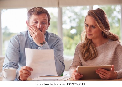 Worried Couple With Bills And Digital Tablet Sitting At Table At Home Reviewing Domestic Finances