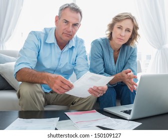 Worried Couple Paying Their Bills Online With Laptop Looking At Camera At Home In The Living Room