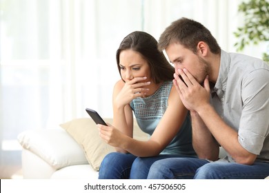 Worried couple reading bad news together on the phone sitting on a sofa in the living room at home
