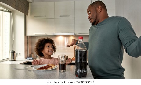 Worried Dad Shrug And Look At Cheerful Little Son Have Lunch Or Dinner With Cola And Pizza At Table At Home Kitchen. Unhealthy Eating. Black Family Lifestyle And Relationship. Fatherhood And Parenting