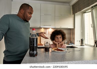 Worried Dad Shrugging Near Offended Son Having Lunch Or Dinner With Cola And Pizza At Table At Home Kitchen. Unhealthy Eating. Young Black Family Lifestyle And Relationship. Fatherhood And Parenting