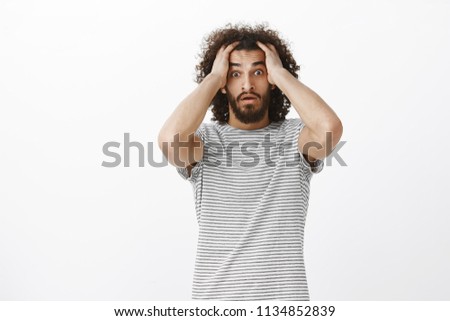 Worried desperate aduly male freelancer with afro haircut in trendy striped t-shirt, holding hands on head and lifting eyebrown, feeling nervous and shocked, seeing work not finished in time